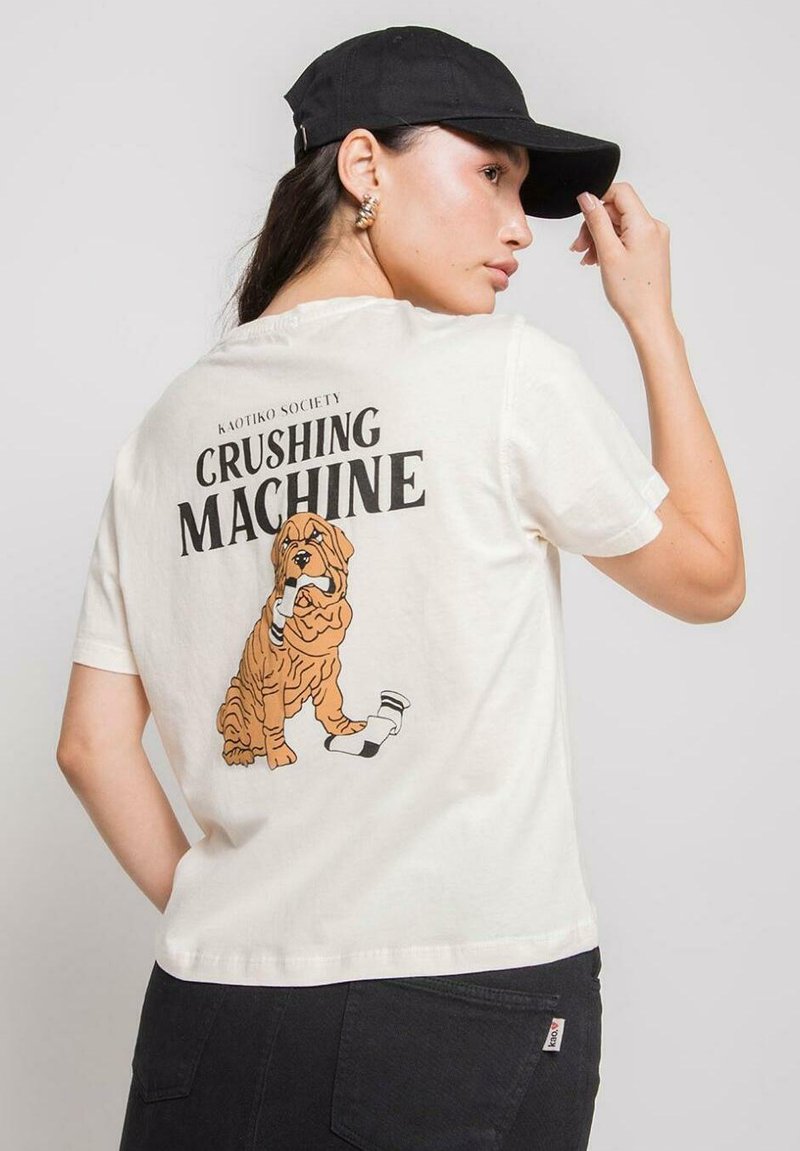 Camiseta blanca de manga corta con el texto "Máquina Aplastante" y un gráfico de un perro marrón sosteniendo un calcetín. Combinada con una gorra negra.