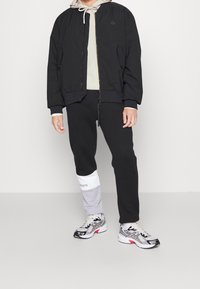 Blouson aviateur noir avec poignets côtelés, sweat à capuche beige en dessous, pantalon de survêtement noir avec une bordure grise portant un logo, et baskets gris et blanc.