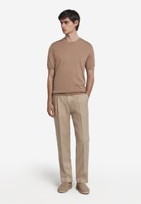 Lysebrunt kortærmet sweater, parret med beige plisserede bukser og lysebrune espadriller, stående mod en hvid baggrund.