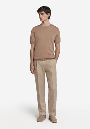 Lysebrunt kortærmet sweater, parret med beige plisserede bukser og lysebrune espadriller, stående mod en hvid baggrund.