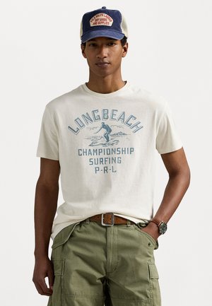 Off-white cotton t-shirt features blue graphic text reading "LONG BEACH CHAMPIONSHIP SURFING P-R-L." Pair of green cargo shorts visible below.