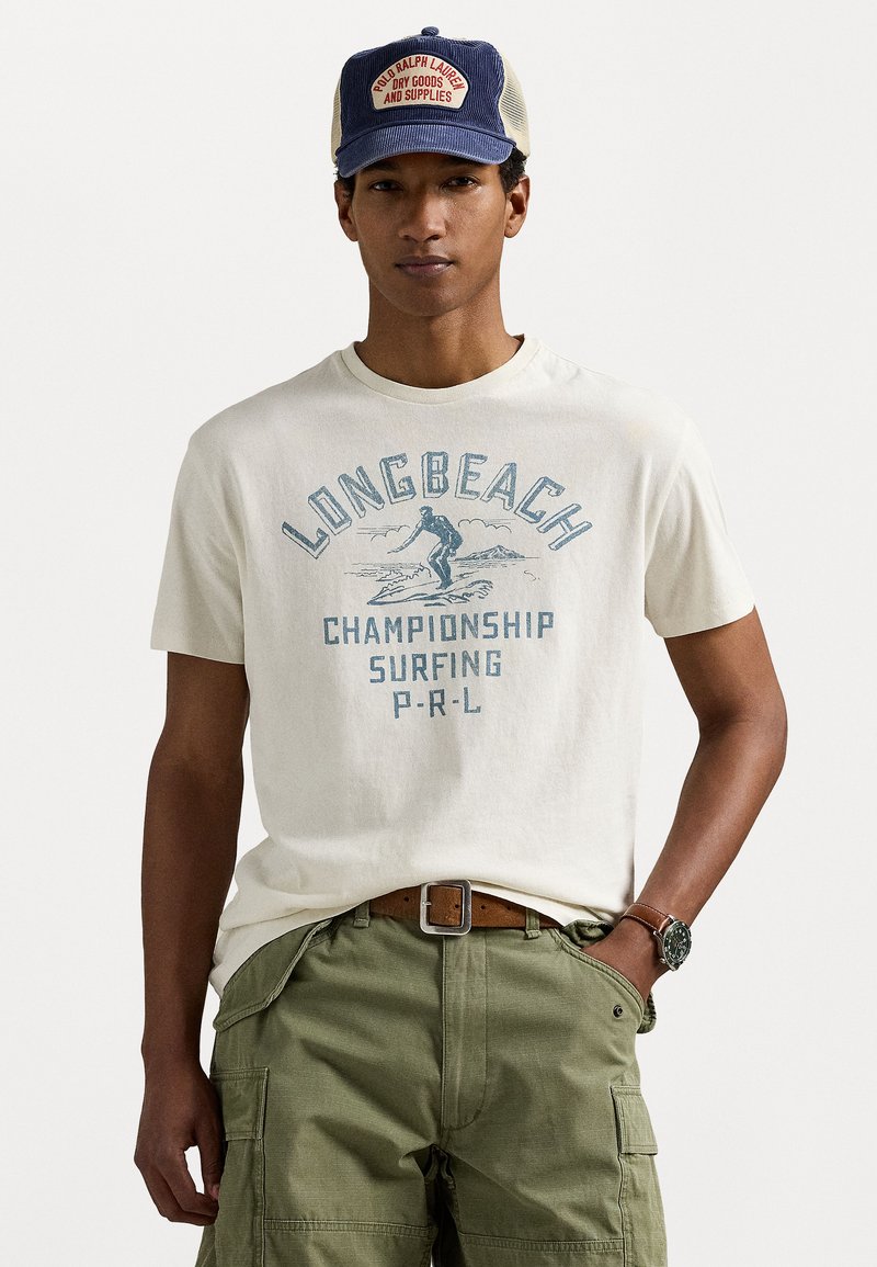 Oficialiai balta medvilninė marškinėliai turi mėlyną grafiką tekstą, kuriame rašoma "LONG BEACH CHAMPIONSHIP SURFING P-R-L." Žemiau matosi žali kargo šortai.