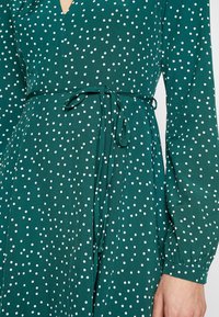 Vestido de color verde azulado con lunares blancos, mangas largas, cinturilla con lazo y escote en forma de V. Tejido suave con una textura lisa.