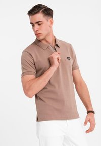 Pánská polokošile s krátkým rukávem z lehkého hnědého bavlněného materiálu, s límcem na zip a malým černým logem na hrudi. Nositelná s bílými kalhotami.