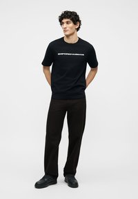 Hombre vistiendo una camiseta negra de Emporio Armani, pantalones oscuros y zapatillas negras, de pie con las manos detrás de la espalda y mirando ligeramente hacia la izquierda.