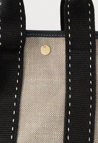 Μπεζ πλεκτή τσάντα tote με μαύρους χοντρούς ιμάντες, με λευκή ραφή και ένα μικρό χρυσό κουμπί σε σχήμα κύκλου.