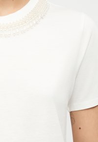 Λευκό πουκάμισο με κοντά μανίκια και διακοσμητικό γιακά με πέρλες, που δείχνει μέρος του βραχίονα με γεωμετρικό τατουάζ και μια μικρή σκοτεινή ελιά κοντά στον αγκώνα.