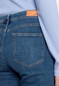 Blue jeans escuros com um corte de cintura alta, apresentando um remendo de logo em couro na cintura e um design clássico de cinco bolsos.
