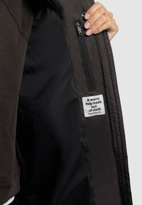 En hand håller upp en mörk jacka som visar en inre ficka med dragkedja och en etikett som säger "En varm kram gjord av tyg."