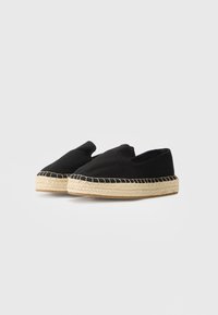 Espadrilles à enfiler en toile noire avec semelles compensées en jute tissé naturel sur une surface blanche.