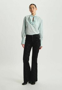 Chemisier bleu clair à volants avec un col en forme de trou de serrure, associé à un pantalon évasé noir à sequins taille haute comportant cinq boutons et une finition texturée.