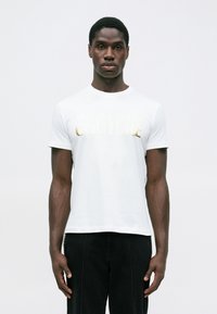 Mužský model stojí na bílém pozadí, oblečený v bílém tričku Versace Jeans Couture se zlatým nápisem a černými kalhotami.