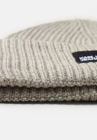 Kootud beanie heledast hallist värvist, millel on tekstuuriga ribimuster. Omab musta sildi valge tekstiga. Materjal näib olevat pehme ja soe.