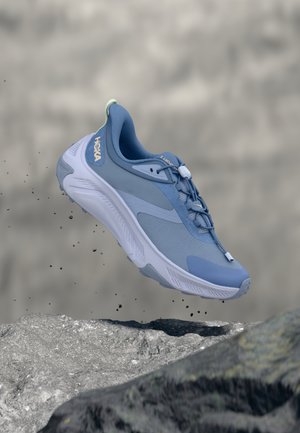 Μπλε και λευκό παπούτσι τρεξίματος Hoka αιωρείται πάνω από πετρώδες έδαφος με μικρά απορρίμματα να πέφτουν γύρω του.