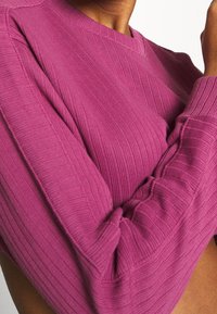 Ribbadsstrickad tröja i livlig rosa med V-halsdesign och voluminösa ärmar. Har en strukturerad tyg och åtsittande mudd för en snug passform.