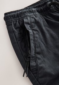 Pantaloni neri con vita elastica, tasca con zip parzialmente aperta e coulisse nere su una superficie liscia e chiara.