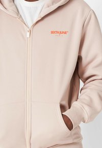 Sudadera de color rosa claro con cremallera, con bolsillos frontales, puños acanalados y un logo naranja de "SIXTH JUNE". Tejido suave con una textura lisa.