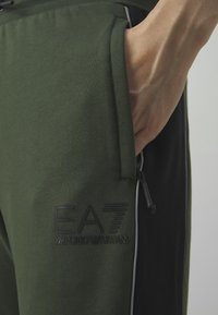 Zelené tepláky s černým logem, bočními kapsami a kontrastním černým panelem. Materiál je měkký a hladký na dotek.