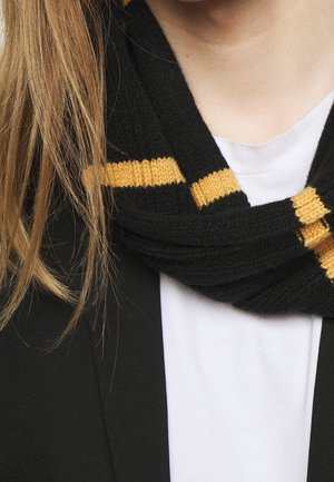 Personne blonde portant un blazer noir, une chemise blanche et une écharpe noire à rayures horizontales jaunes autour du cou.