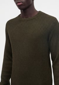 Homme portant un pull en maille vert olive à manches longues avec un col rond sur un fond uni.