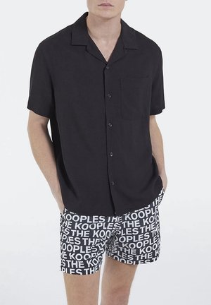 Mężczyzna w czarnej koszuli z krótkim rękawem zapinanej na guziki oraz czarno-białych szortach z powtarzającym się wzorem tekstu "THE KOOPLES".