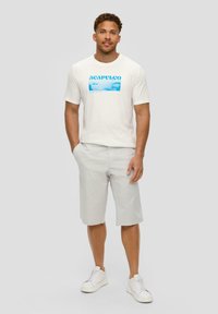 T-shirt blanche avec un graphique bleu "ACAPULCO", associée à un short gris clair. Le modèle porte des baskets blanches ; manches courtes et coupe décontractée.
