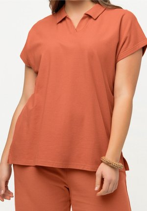 Femme portant un haut ample à manches courtes de couleur rouille et un pantalon assorti, avec un bracelet tressé au poignet droit.