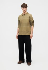 Νεαρός άνδρας φορούμενος ελιάς μακρυμάνικο πόλο πουκάμισο, μαύρο παντελόνι με φαρδιές μπατζάκια και καφέ παπούτσια, στέκεται μπροστά σε απλό λευκό φόντο.