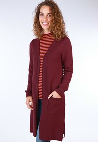 Burgundy long cardigan with a textured pattern, open front, and two pockets, paired with a striped turtleneck and blue jeans.
