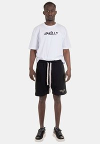 T-shirt bianco a maniche corte con logo "GAËLLE", abbinato a pantaloni corti neri con lacci crema e nome del marchio ricamato. Sneakers nere.