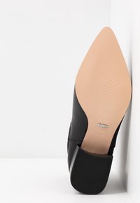 Chaussure à talon haut en cuir noir avec un bout pointu, un panneau latéral noir texturé et une semelle en caoutchouc beige. Présente une gravure de la marque sur la semelle.