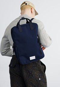 Tummansininen reppu, joka on valmistettu kankaasta ja jossa on kaksi yläkahvaa sekä sivuvetoketjut. Siinä on suorakaiteen muotoinen, minimalistinen muotoilu ja etiketti.