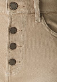 Närbild av beige denimtyg med fyra metallknappar märkta "Marc O'Polo DENIM" och synlig sömnad nära en ficka.