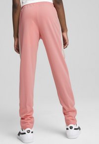 Roza športna hlača z gladko teksturo, elastičnim pasom in kontrastnimi stranskimi trakovi. Obute so v bele in črne superge.