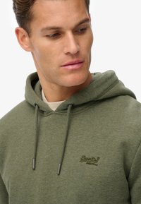 Oliven grøn hoodie lavet af tekstureret stof med snoretræk i huen, der har et broderet logo på brystet i matchende tråd.