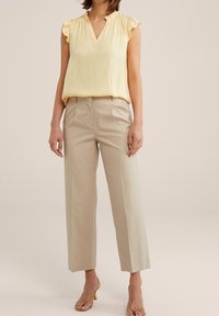 Femme portant un haut sans manches jaune pâle avec des épaules volantes, un pantalon taille haute beige et des sandales à talons beiges sur un fond neutre.