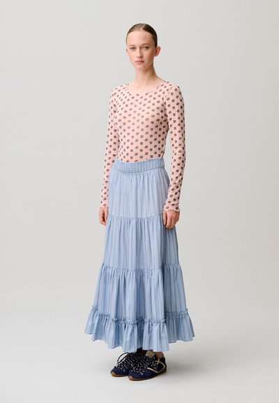 Modella in piedi che indossa una maglia floreale a maniche lunghe, una gonna lunga azzurro chiaro a balze e sneakers blu navy su sfondo neutro.