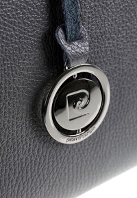 Primo piano di un ciondolo rotondo argentato con logo a spirale attaccato a una tracolla in pelle nera testurizzata.