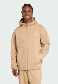 Beige színű, cipzáras kapucnis pulóver, textúrált felülettel, kapucnival és oldalsó zsebekkel. Diszkrét Adidas logó található a mellkason és az ujjon.