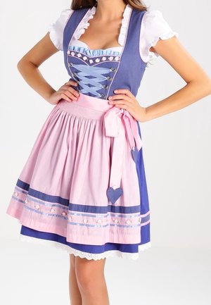 Kvinde iført en traditionel blå og lyserød dirndl-kjole med en hvid bluse, lyserødt forklæde, bånddetaljer og hænderne i siden.