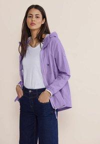 Letvægts, lilla vindjakke med hætte, frontlynlås og sidelommer. Bæret over en hvid skjorte og mørke jeans. Glat tekstur.