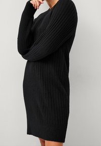 Robe en maille noire côtelée avec manches longues, encolure ronde et taille cintrée. La texture est douce avec une légère élasticité.