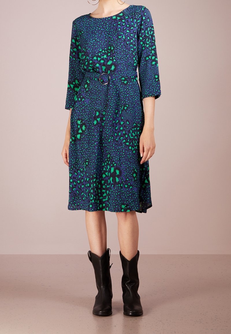 Femme portant une robe bleu et verte à motifs, longueur genou, manches 3/4, et des bottes noires mi-mollet, debout sur un sol uni.