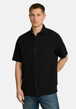 Young man wearing a black short-sleeve button-up shirt and blue jeans standing against a plain light background.