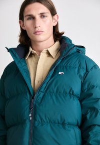 Teal puffer jacket with a zip front and a hood. Features stitched sections and a small logo patch. Worn over a beige corduroy shirt.