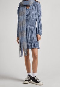 Sininen kukkamekko pitkillä hihoilla, elastinen vyötärö, röyhelöhelma, yhdistettynä vaaleansinisellä ja harmaalla ruutuhuivilla sekä mustilla lenkkareilla.