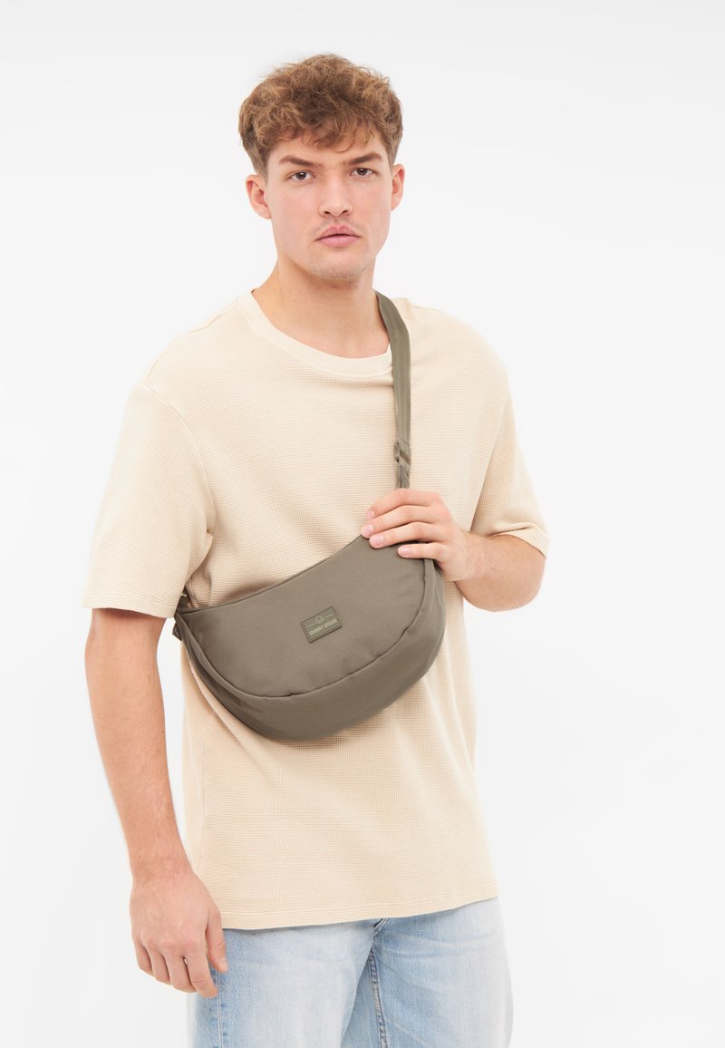 Borsa a tracolla verde oliva, tessuto liscio, forma ovale, chiusura con zip, patch logo frontale, tracolla regolabile, indossata sopra una camicia beige con texture.