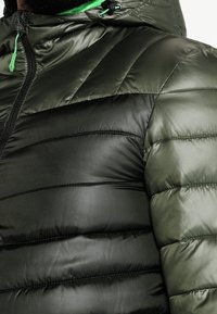 Tumma vihreä untuvatakki kiiltävällä pinnalla, jossa on vaakasuorat ompeleet, vetoketjullinen etuosa ja naruilla säädettävä huppu vihreillä yksityiskohdilla.