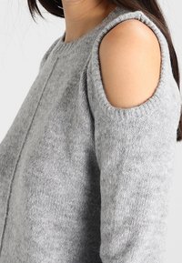 Grau gestrickter Pullover mit rundem Ausschnitt und Cutouts an den Schultern. Der Stoff hat eine weiche Textur mit einem dezenten Muster.