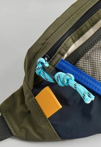 Olivgrön väska med svart dragkedja, blå detaljer, nätficka och en gul tyglapp. Texturerad yta och justerbar blå snodd.